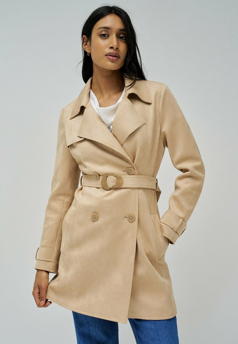 Trench-coat beige à double boutonnage avec une taille ceinture. Fabriqué en tissu doux et texturé, avec de grands boutons et un col à revers.