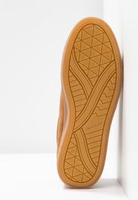 Semelle d'une chaussure en cuir suédé marron avec une semelle extérieure en caoutchouc à motifs, le nom de la marque "GANT" et la taille "37" visibles, inclinée contre une surface blanche.