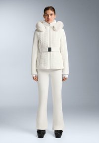 Giacca bianca con cappuccio foderato in pelliccia, design a maglia intrecciata testurizzata, vita con cintura. Abbinata a pantaloni bianchi a zampa e scarpe con platform nere.