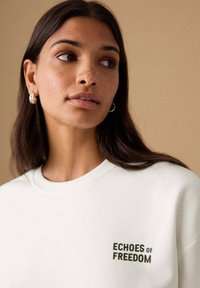 Hanorac alb cu broderie "ECHOES OF FREEDOM" în verde închis. Prezintă decolteu rotund și manșete striate. Textură netedă a țesăturii.