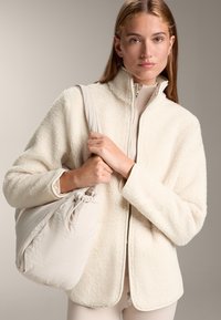 Giacca in pile color crema con colletto alto e chiusura con zip. Il modello tiene una borsa slouchy in tessuto morbido di un leggero beige. Sfondo neutro.