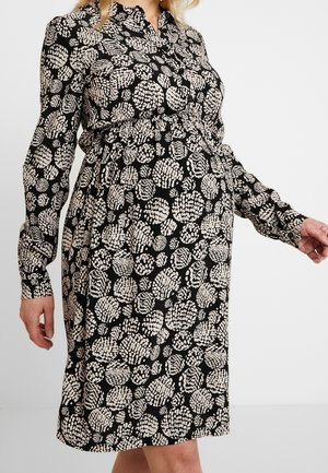 Schwangere Frau trägt ein schwarzes langärmliges Kleid mit beigen abstrakten Kreismustern und steht vor einem schlichten Hintergrund.