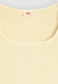 Κίτρινη ριμπ τοπ με φαρδύ ντεκολτέ, με ετικέτα με κόκκινο κείμενο. Το ύφασμα φαίνεται μαλακό και ελαφρώς υφασμάτινο.