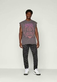 Ärmelloses graues Grafik-T-Shirt mit lila Druckdesign, kombiniert mit schwarzen gemusterten Hosen und weißen Sportschuhen.