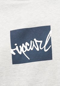 Λευκό καλλιγραφικό κείμενο «ripcurl» επάνω σε ναυτικό μπλε τετράγωνο σε ανοιχτό γκρι υφασμάτινο φόντο.