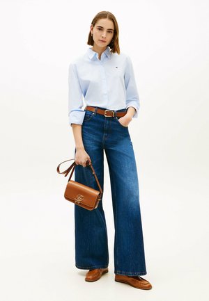 Ung kvinde iført en lyseblå, stribet skjorte, vide jeans, brunt bælte og sko, der holder en brun læderhåndtaske i remmen.
