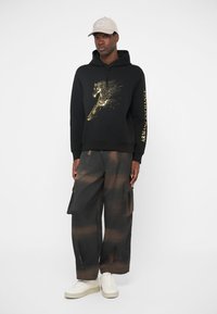 Μαύρη κουκούλα με χρυσό σχέδιο αλόγου και κείμενο "ARMANI EXCHANGE". Συνδυασμένη με παντελόνια cargo με καφέ ρίγες και λευκά αθλητικά παπούτσια.