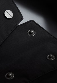 Tessuto di camicia nera con una sottile texture, che presenta due bottoni a pressione metallici e un bottone argento con logo. Dettaglio ravvicinato del colletto.