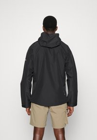 Veste noire imperméable avec capuche, manches longues et poignets élastiques ; portée par-dessus un short beige ; présente une texture lisse et un design minimal.