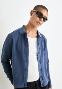 Blue long-sleeve button-up shirt with a collar. Worn over a white ribbed tank top. Black angular sunglasses complete the outfit.