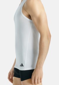 Bílý sportovní tílko s upnutým střihem, vyrobené z hladkého materiálu, s černým logem Adidas a akcentovými pruhy na bocích.