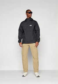 Giacca impermeabile nera con cappuccio e logo bianco, abbinata a pantaloni cargo beige e sneakers bianche. Occhiali da sole completano il look.