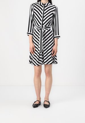 Femme portant une robe boutonnée à rayures diagonales noires et blanches avec ceinture à nouer à la taille et manches trois-quarts, associée à des ballerines noires.