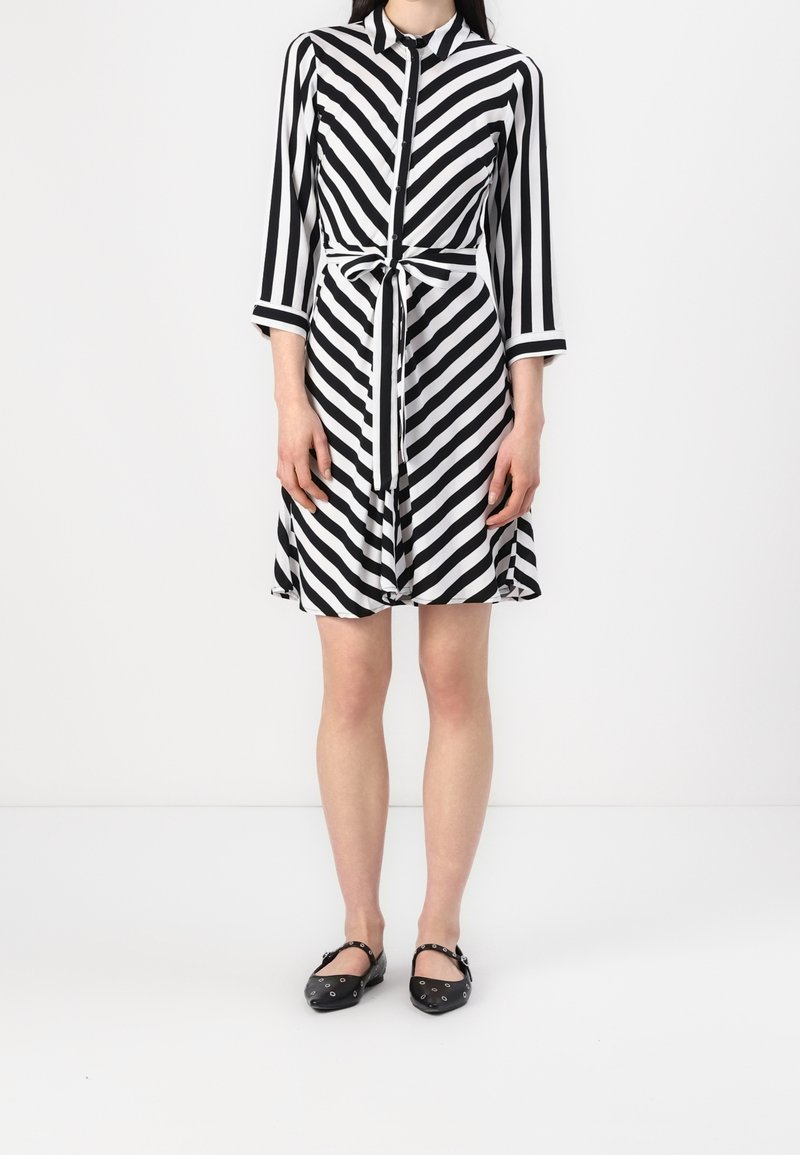 Femme portant une robe boutonnée à rayures diagonales noires et blanches avec ceinture à nouer à la taille et manches trois-quarts, associée à des ballerines noires.