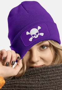 Purple knit beanie featuring a white skull and crossbones graphic on a rectangular patch; ribbed texture with a snug fit.