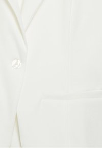 Gros plan sur le tissu d'un blazer blanc montrant un seul bouton blanc, le revers et une poche passepoilée à l'avant.