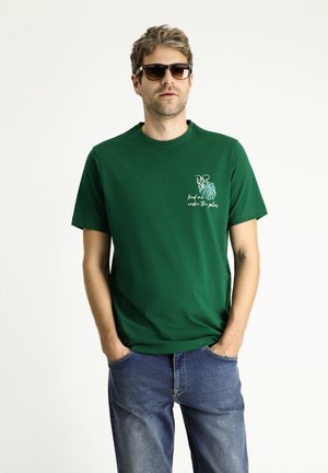 Groene t-shirt met korte mouwen en een ronde halslijn, voorzien van een grafische print aan de voorkant met tekst en een palmbladontwerp. Draagt met blauwe spijkerbroek.