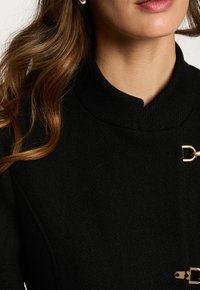 Abrigo negro con un collar alto, tela texturizada y acentos de hardware dorado. Presenta un cierre con un diseño distintivo.