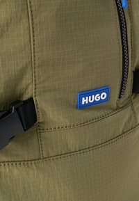 Tkanina w oliwkowym kolorze z teksturą kratki, z czarnym zamkiem błyskawicznym, niebiesko-białą naszywką z logo "HUGO" i czarnymi elementami metalowymi na pasku.