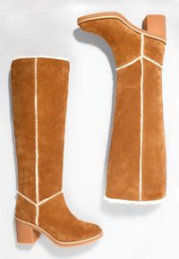 Bottes montantes pour femmes en daim beige avec coutures blanches visibles, bouts arrondis, talons épais et doublure en fausse fourrure le long des bords.