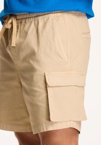 Pantaloni scurți cargo bej cu talie elastică, închidere cu șnur, buzunare laterale și un buzunar cu clapetă pe coapsă. Material ușor.