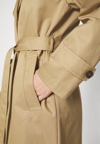 Trench coat beige avec taille ceinturée et lanière au poignet, main placée dans la poche latérale.