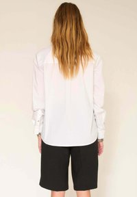 Chemise blanche à manches longues avec des poignets à boutons, associée à un short noir. La chemise présente un ourlet arrondi et un design simple.