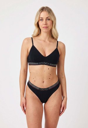 Donna con capelli biondi che indossa un reggiseno nero e slip a vita alta abbinati con fasce elastiche a righe arcobaleno e logo.