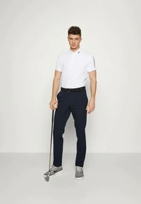 Camisa polo blanca con cuello y logotipo lateral, combinada con pantalones navy. Zapatillas grises y un palo de golf acentúan el look deportivo.