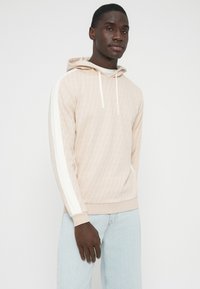 Bež pulover s kapuco s teksturiranim vzorcem diamantov in belimi poudarki na rokavih. Ima vrvice za zategovanje in sproščen kroj, kombiniran s svetlimi kavbojkami.