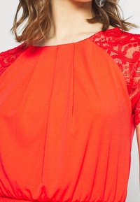 Czerwona sukienka z plisowanym wzorem i krótkimi koronkowymi rękawami. Tkanina wydaje się gładka, z lekko marszczoną talią dla lepszego dopasowania.