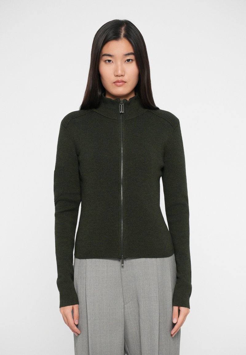 Σκούρο πράσινο φούτερ με φερμουάρ και ψηλό γιακά, ριμπ λεπτομέρειες και μακριά εφαρμοστά μανίκια, συνδυασμένο με ανοιχτό γκρι παντελόνι με φαρδιά πόδια.