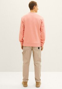 Różowa bluza z okrągłym dekoltem i ściągaczami, w połączeniu z jasnobeżowymi spodniami cargo z kieszeniami. Stylizacja jest teksturalna i swobodna.