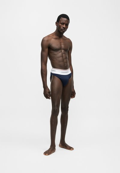 Αθλητικός άνδρας στέκεται ξυπόλυτος μπροστά σε λευκό φόντο, φορώντας σκούρα μπλε εσώρουχα με λευκή μέση, στραμμένος ελαφρώς αριστερά με σοβαρή έκφραση.