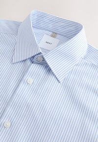 Chemise blanche avec fines rayures verticales bleues, col pointu, boutons blancs et une étiquette indiquant NEXT à l’intérieur du col.