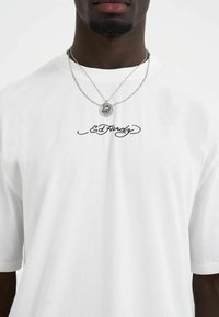 Hvid kortærmet t-shirt med sort skråskrift "Ed Hardy" logo. Laget sølvkæder med rund vedhængs halskæde. Ensfarvet baggrund.