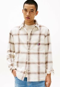 Button-up plaid shirt in white, brown, and blue tones, featuring a chest pocket and a relaxed fit. Made from soft fabric with a textured surface.