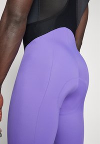 Lila sportleggings sima felülettel, kontúros szabású, kontrasztos fekete felső résszel, részletes varrásokkal és testhezálló kialakítással.