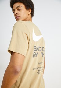 Bēšs Nike T-krekls ar baltu Nike swoosh logotipu un grafiku uz muguras, īsām piedurknēm un gludu kokvilnas tekstūru.