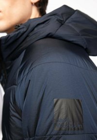 Granatowa kurtka puchowa o fakturowanym wykończeniu, z kapturem i czarną naszywką z logo na lewym ramieniu.