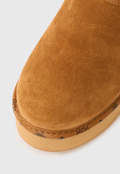 Bottine en daim marron avec un bout arrondi, une semelle extérieure contrastante couleur tan, et un branding discret le long du bord. Texture lisse, design minimaliste.