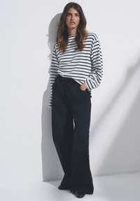 Camisa de manga larga a rayas en blanco y azul marino, combinada con pantalones negros holgados. El conjunto incluye una cintura con cordón y un corte relajado.