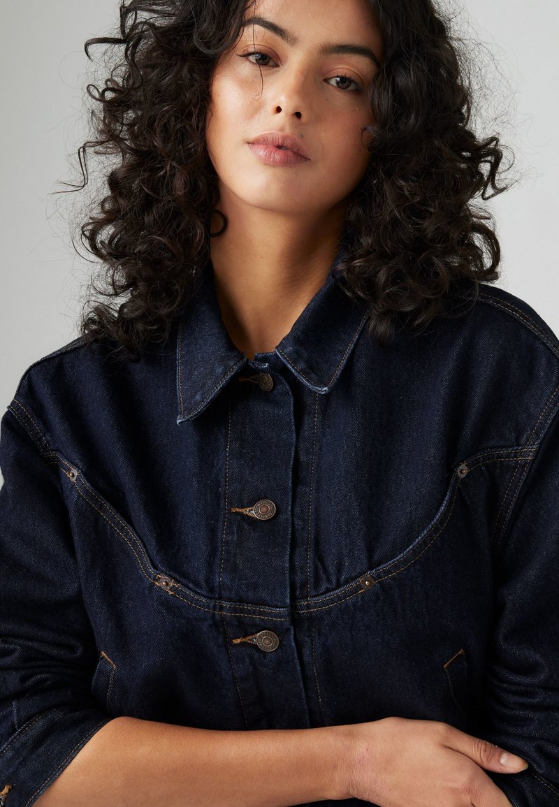 Giubbotto di jeans blu scuro con colletto, bottoni frontali e cuciture visibili. Presenta tasche laterali e una vestibilità leggermente ampia.