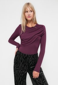 Body à manches longues en violet profond avec un design drapé sur le devant, associé à un pantalon noir avec un motif texturé subtil.