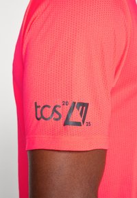 Tricou sportiv de culoare roz strălucitor, cu o textură din plasă, având un logo imprimat în negru pe mânecă care afișează "tcs 20 25".