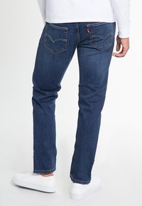 Man i mörkblå jeans och vita sneakers som står mot en enfärgad bakgrund, sett bakifrån.