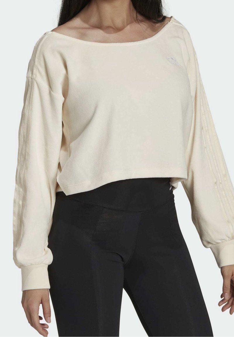 Krämfärgad kort sweatshirt med vid halsringning och långa, texturerade ärmar, kombinerad med svarta leggings. Enkel branding på bröstet.