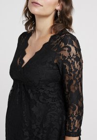 Robe en dentelle noire avec des manches longues, un col en dentelle festonné, des motifs floraux et un tissu texturé. Détails de manches semi-transparentes.