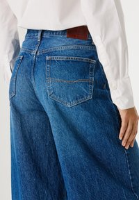 Wide-leg blue denim jeans feature a high waist, standard back pockets, and prominent orange stitching on the seams. Leather branding on the waistband.
