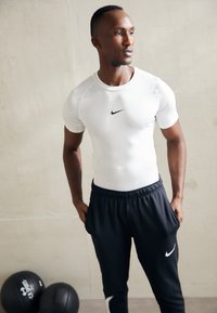 Camisa atlética blanca de corte ajustado para hombres con mangas cortas, que presenta un logo negro de Nike, combinada con pantalones de chándal azul marino oscuro con bolsillos laterales.